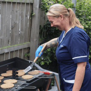 Woman outside in a Dale Care uniform wearing blue latex cloves and flipping burgers on a BBQ.