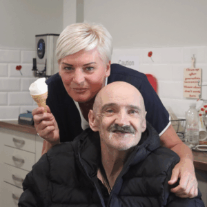 A Dale Care carer with a service user, both are smiling. The carer is holding an ice cream in one hand and has her other hand on the service user's shoulder.