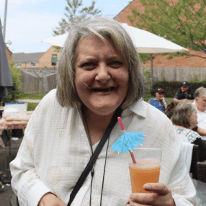 An older woman with grey hair and a cream button up top standing outside. She is smiling at the camera and holding a mocktail with a drink umbrella in it.