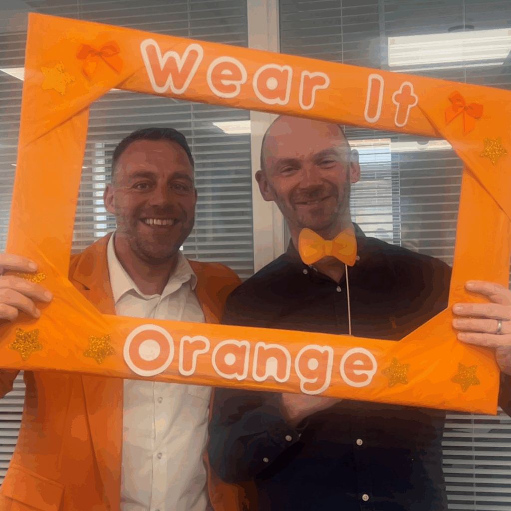 Two men, one wearing an orange jacket and the other an orange bowtie holding a sign that says 'Wear It Orange'.