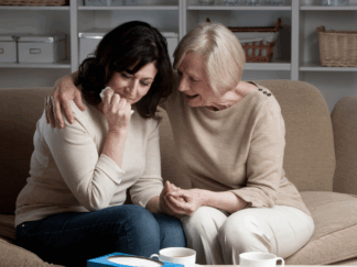 Two women sitting in a living room wearing beige coloured clothing. The woman one the left looks upset and is clutching a tissue while the woman on the right comforts her.