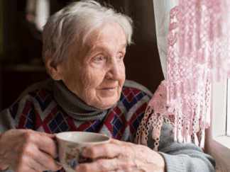 A lonely elderly individual holding a beverage, looking out of the window at Christmas.
