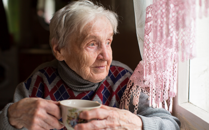 A lonely elderly individual holding a beverage, looking out of the window at Christmas.