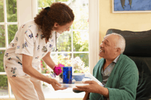 Carer interacting with a service user and handing him a tray with food and drink while he sits on a chair wearing a dark green dressing gown.