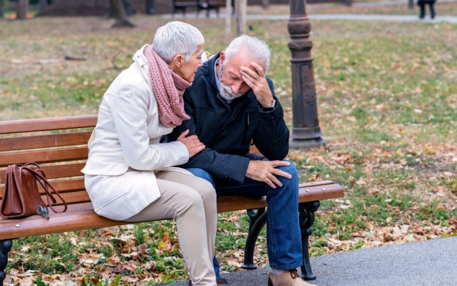 Depression in older adults representation: Two older adults sitting on a park bench outside. One man and one woman, presumably a married couple. Man has his head in his hands, looking distressed and woman is comforting him.