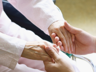 Two hands gently grasping another person's hands. While faces can't be seen, one pair of hands looks older and you can see that this person is sat down.