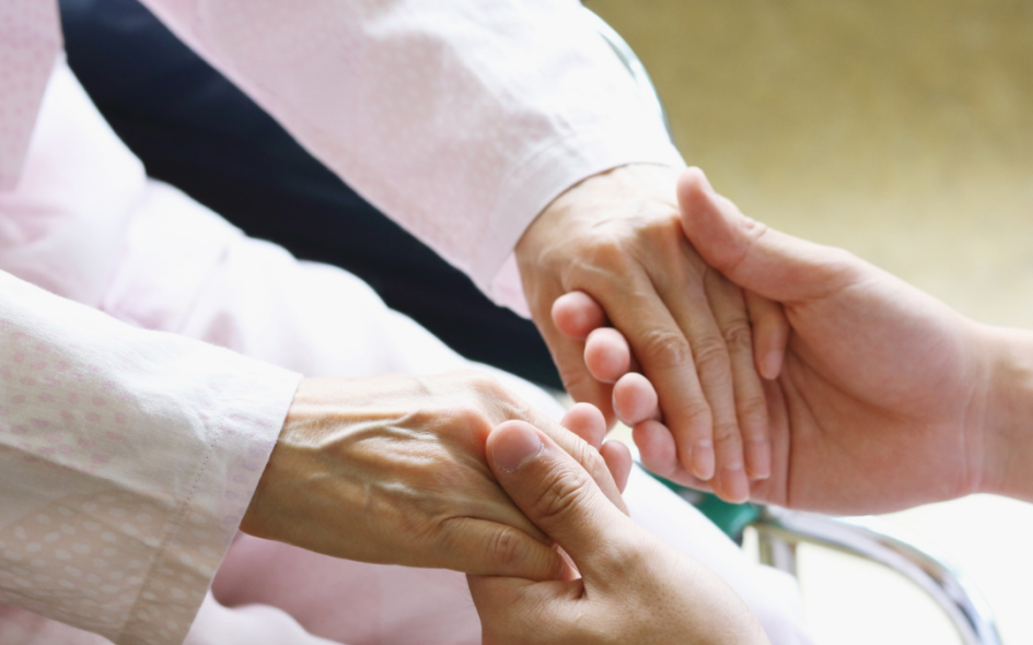 Two hands gently grasping another person's hands. While faces can't be seen, one pair of hands looks older and you can see that this person is sat down.